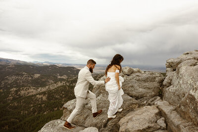 groom helps bride over rocks as they hike to their elopement ceremony in Boulder Colorado.