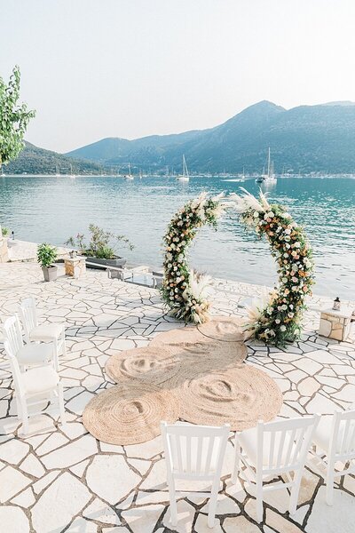 Romantic wedding ceremony setup featuring a circular floral arch with pampas grass and roses, overlooking the water and anchored by woven jute rugs