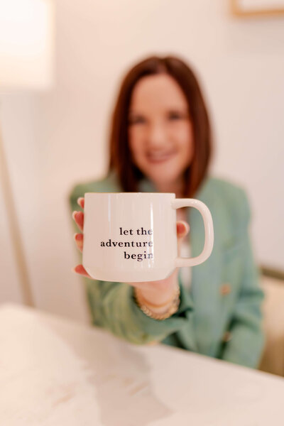 Woman wearing green blazer holding mug that reads "Let the Adventure Begin" toward camera