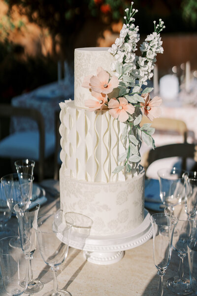 All white 3-teir wedding cake with curved texture design on middle tier and stencil design on top and bottom tiers and peach and white sugar flowers