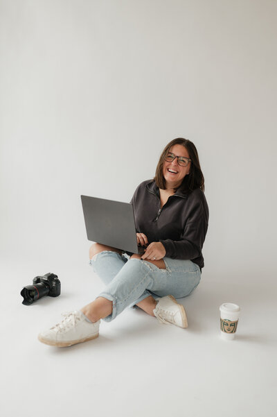 A person smiling as they sit on the floor with a lap with a camera and coffee cup next to them 