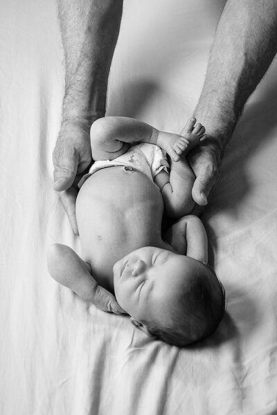 A black and white photo of a newborn baby lying on a bed, with large, gentle hands cradling the baby’s sides, emphasizing the tenderness and care of the moment.