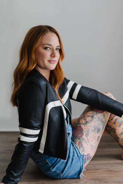 Woman wearing a black leather jacket and jean shorts sits on the floor inside a North Carolina photo studio | Sara Coffin Photo
