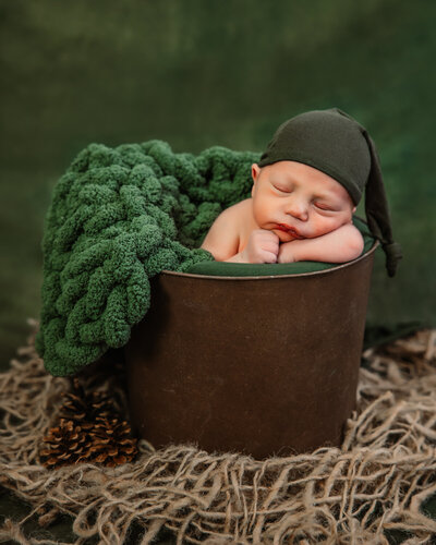 newborn boy in a brown metal bucket with a green blanket and green hat on sleeping peacefully