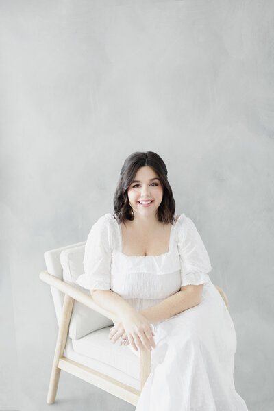 A woman with dark hair in a white dress sits in a chair in front of a grey photography background smiling at the camera.