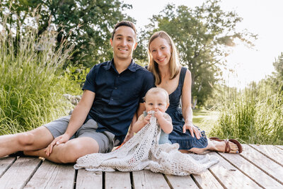 A family of three sits on a wooden deck in a sunlit garden. The father and mother smile at the camera while their baby, wrapped in a crochet blanket, looks up curiously.