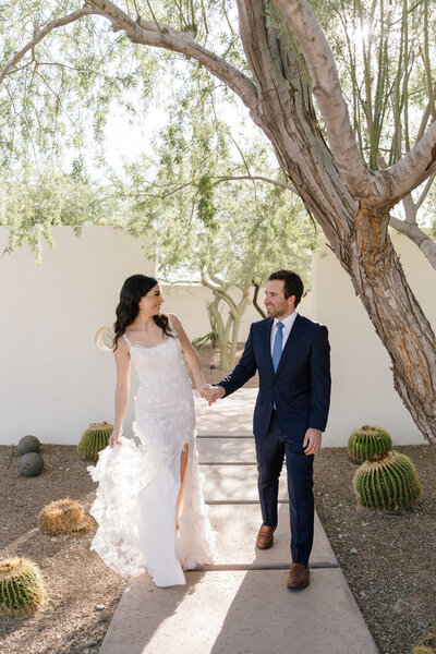 Groom wearing blue suit holding bride's hand walking on paved path and smiling during backyard wedding in Paradise Valley, Arizona.