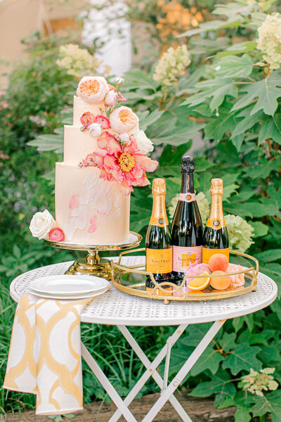 A pink cake with large pink flowers on a table with champagne.
