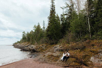 bride and groom sitting on rocks with Lake superior in the background and tall, green, pine trees