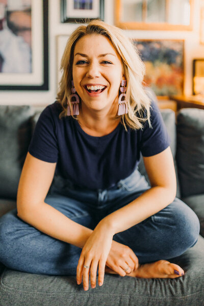 A woman wearing a t-shirt and jeans is laughing while sitting cross legged on a grey couch.