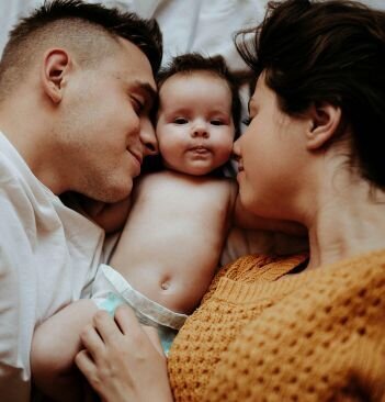 Two parents lying on a bed, with a baby between their heads.