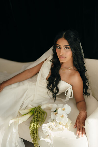 A bride in an elegant white dress reclines on a white couch, holding a bouquet of white orchids and greenery. Her serene expression adds to the calm ambiance.