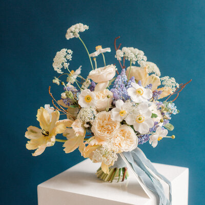A bridal bouquet with white, lilac, and pale peach flowers, standing on a table. 