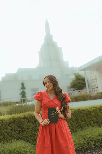 A soon-to-be missionary smiles as she holds a Book of Mormon. She's standing in front of the LDS Idaho Falls temple.