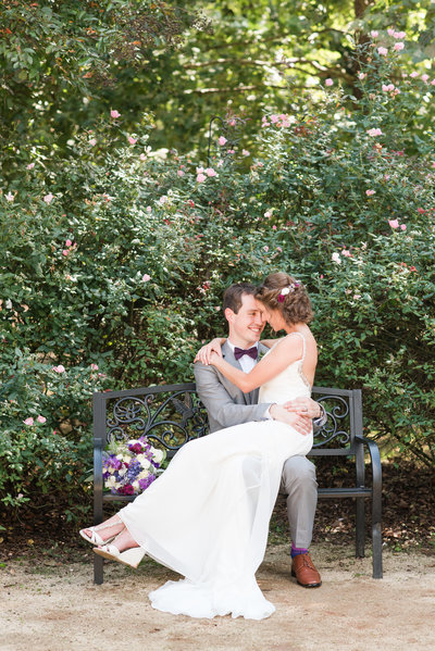 bride sitting on top of groom wearing gray suite with purple bowtie sitting on a bench surrounded by pink roses at Merry Hill Weddings