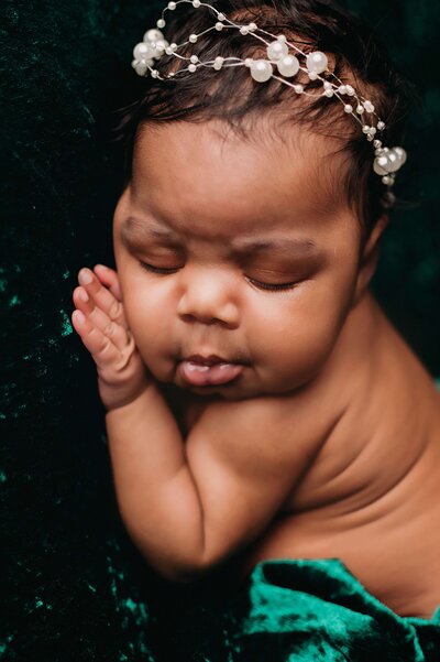 A newborn girl, sleeping with her lips poking out and her hand tucked comfortably under her cheek. She is sleeping on a velvet green backdrop, and is wearing a pearl headband. This detailed newborn photo was captured by Mobile, AL Newborn Photographer