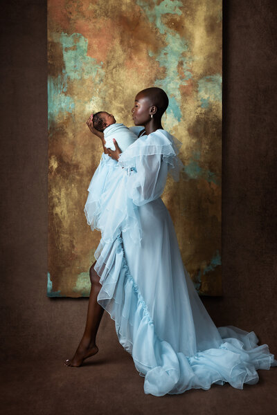 Mother wearing a flowing pale blue gown holding her swaddled newborn baby in front of a gold and teal textured backdrop. Captured by a Dallas Newborn Photographer specializing in luxury motherhood portraits.