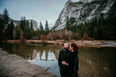 Couple dressed in black attire stands beside a reflective river during their winter elopement in Yosemite National Park, surrounded by pine trees and granite cliffs.