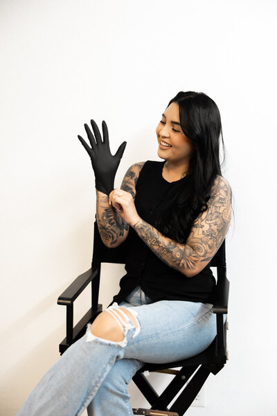 Tattooed hairstylist smiling while putting on black gloves in salon chair. Photograph by Yucaipa branding photographer Kaitlyn Dawn Photography.