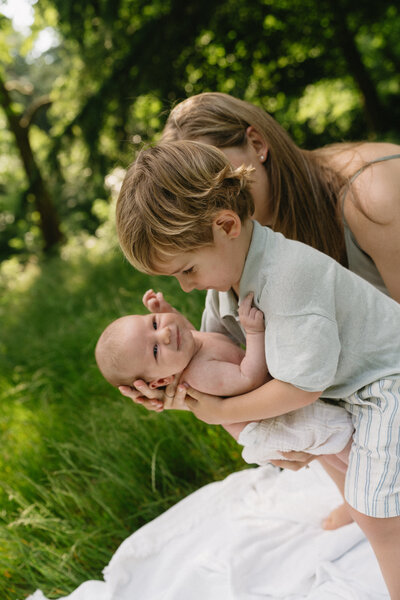 Moeder geeft haar newborn baby aan zoontje dat ernaast staat buiten op een dekentje in het park.