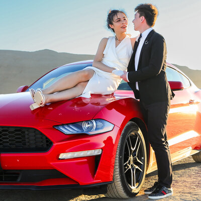 Bride sitting on hood of red mustang  groom standing next to the car embracing the bride