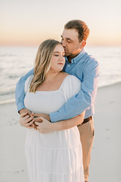 A couple stands on the beach at sunset for their engagement photos.