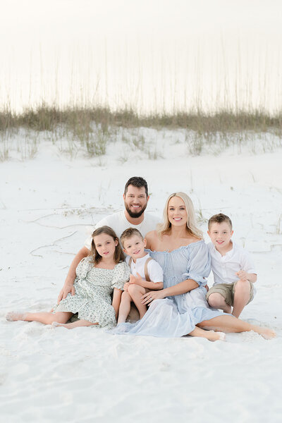A family of five poses barefoot in soft pastel tones on the white sand, with sea oats swaying in the background at golden hour.