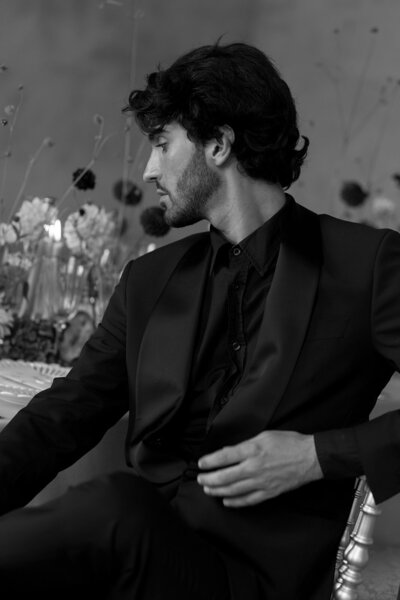 A black-and-white portrait of a groom in a sleek black suit seated elegantly with a floral backdrop.