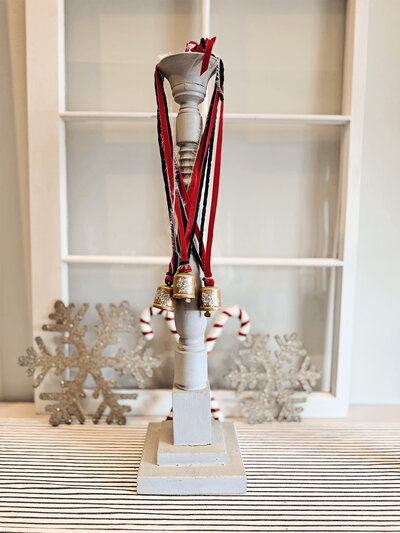 A white candlestick holds several bells with red and green ribbons. Silver glitter snowflakes, a holiday ornament, and two candy canes from Sweet Pea Ink Creative are displayed in the background.