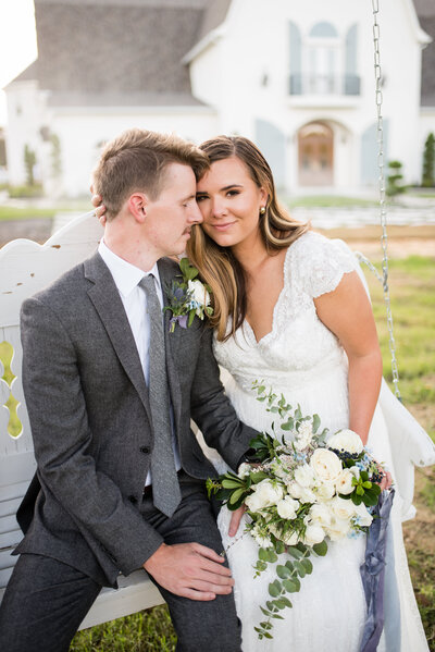 A bride and groom posed with their heads touching while sitting on a bench swing in front of the The French Farmhouse Venue in Collinsville, Texas.