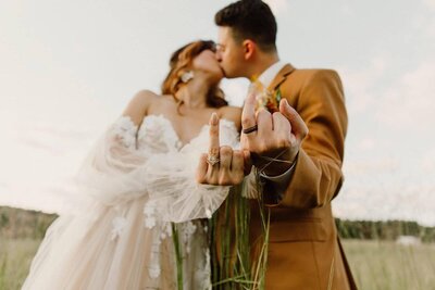 Romantic moment captured in nature as the bride and groom tenderly kiss, showcasing their wedding rings to the camera, symbolizing eternal love and commitment