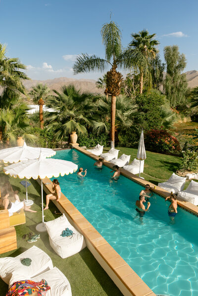 Wedding guests lounge and swim in a pool at a luxury wedding estate in Palm Desert. The pool sparkles and is framed with vintage umbrellas.