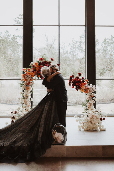 Man in woman in a long black dress kissing in front of a curved floral arrangement of red, orange and white