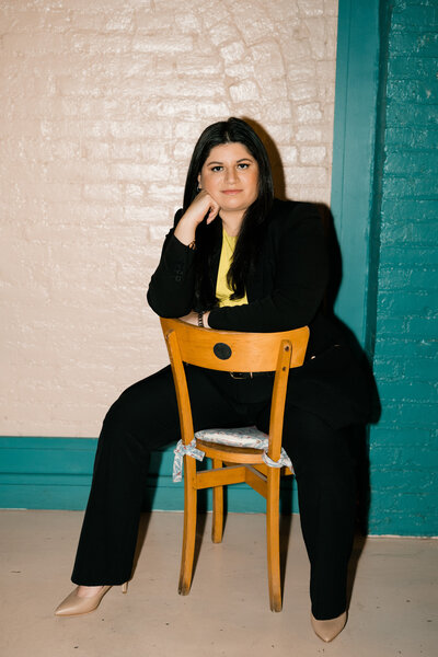 women in a black suit sitting on a wooden chair backwards. while one hand is resting below her cheek
