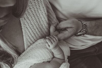 A close-up in soft monochrome of a newborn baby's tiny hand gripping their father's finger, while the mother lovingly looks on. Tender family momen