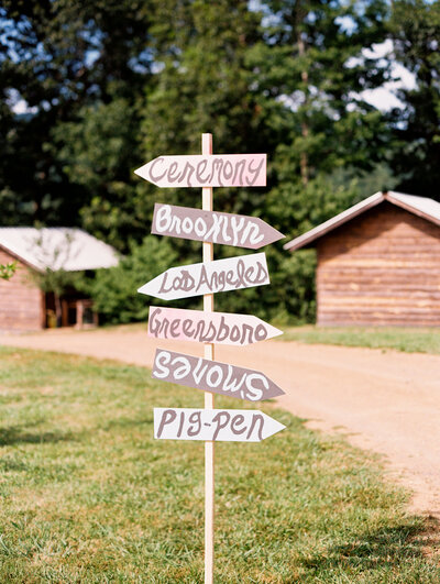 A sign points the directions of important places for the bridal couple, such as the ceremony site, Brooklyn, Los Angeles, and Greensboro, as the ceremony takes place at the wedding venue Paint Rock Farm in North Carolina, by photographer Megan Lynn of My Sun and Stars Co. With cabins in the background.