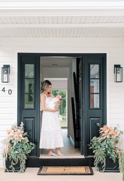 A woman stands in front of her home holding her newborn during her portrait photos in New Jersey