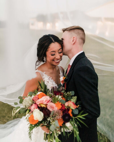 Bride and groom share a joyful, intimate moment, with the bride smiling and holding a colorful bouquet as her veil billows around them.