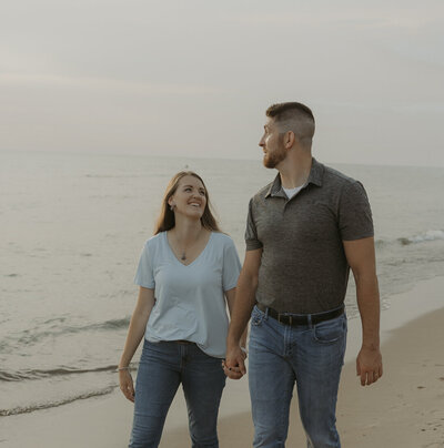 A photograph of a husband and wife walking woodlawn beach, located in western new york, at golden hour while holding hands. the lake to their right, and the beautiful cotton-candy sky behind them. the couples is laughing. Photo by Dash and Boheme Photography. 