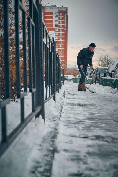 Man in black coat doing snow removal and shoveling sidewalks in front of commercial building