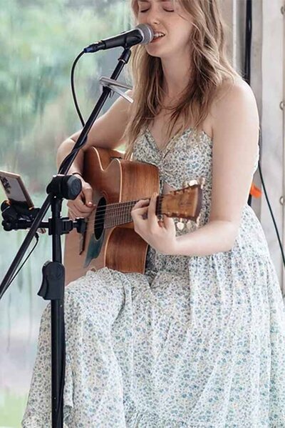 Female acoustic guitarist and vocalist performing outdoors in a floral dress with eyes closed in song.