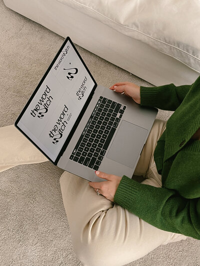 Margarita holds an open laptop with variations of the word witch logo, while sitting on a white carpet floor in white pants and a mossy green sweater