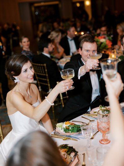 bride and groom sit at table at reception and raise glasses