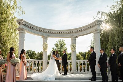 The bride and groom stand beneath a grand stone colonnade, hands clasped as they exchange vows. Bridesmaids in blush gowns hold delicate bouquets, while groomsmen in black tuxedos stand in crisp formation. The soft golden sunlight filters through weeping willow trees, casting a dreamy glow over this elegant wedding ceremony