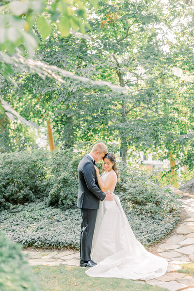 A Portrait of a bride and groom holding each other close standing under the trees on a garden path in Washington DC