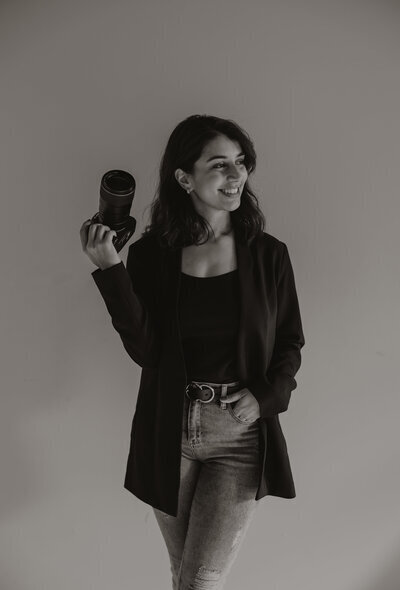 A woman in a black top and blazer smiles while holding a camera, embodying her role as a New England Lifestyle Photographer. With one hand in her pocket against a neutral background, she exudes professionalism. The black and white image captures the essence of timeless artistry.