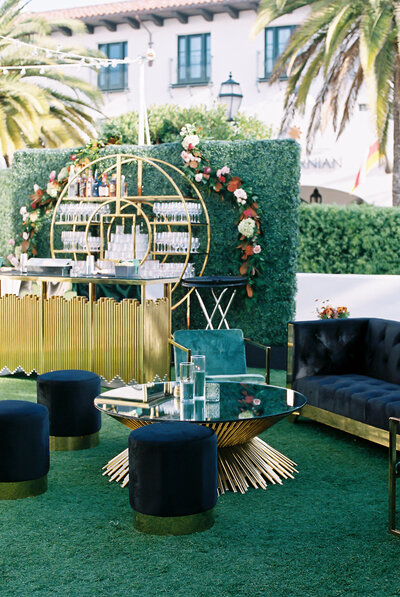 Luxury outdoor event lounge by BP Collective featuring a gold circular bar with champagne display, velvet seating, floral wall backdrop, and modern glass tables.