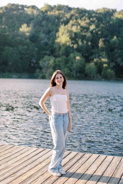 senior girl with one hand on her hip, standing on a dock by a lake