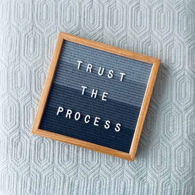 Image shows a grey framed letterboard with the phrase 'Trust the Process' spelled out in white letters. The letterboard is placed on a textured light blue background