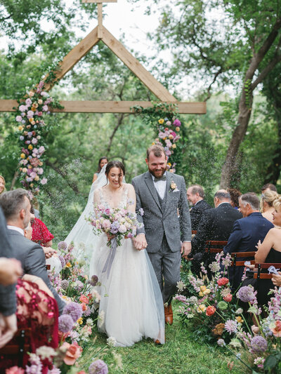 A bride and groom walks down the aisle holding an Anthousai bouquet and flowers at the base of the aisle.